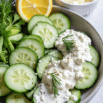 Sliced cucumbers in a ceramic bowl topped with tzatziki, lemon, dill, and crumbled feta on the side.