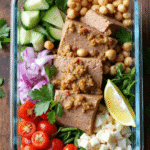 Meal prep glass container filled with protein-packed tuna garbanzo bean salad, featuring sliced cucumbers, chickpeas, red onion, cherry tomatoes, parsley, white cheese cubes, and a lemon wedge.
