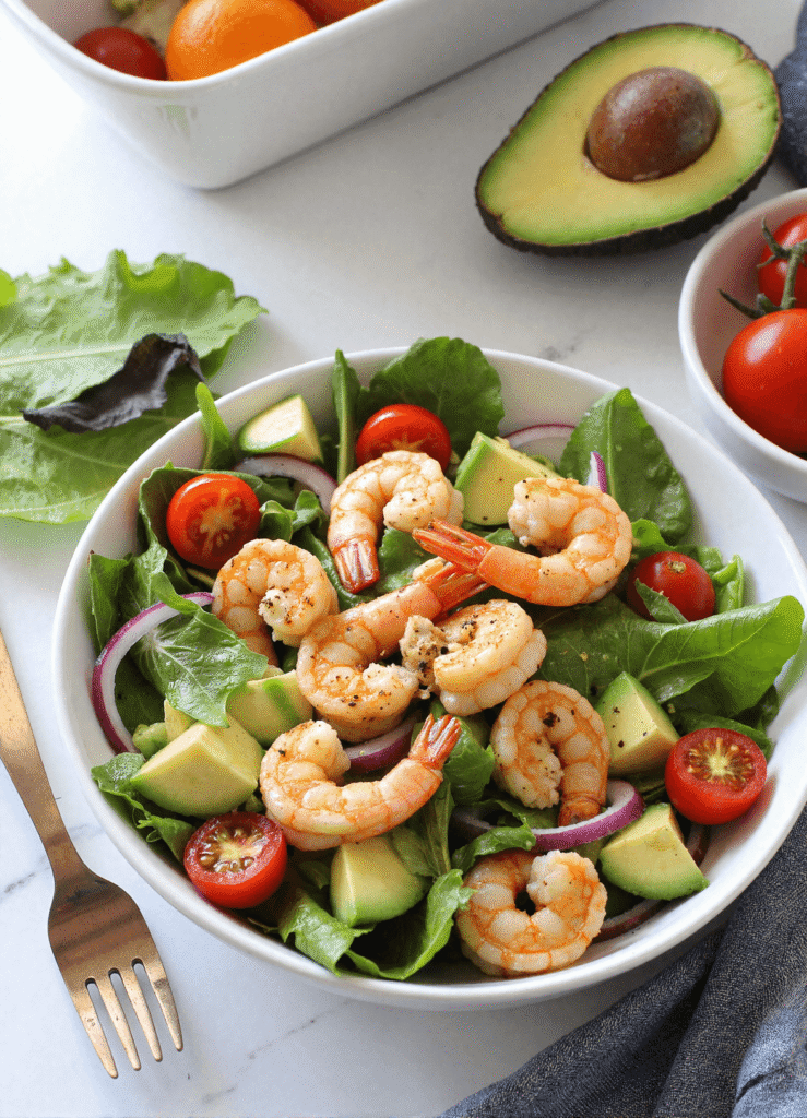 shrimp and avocado salad with cherry tomatoes, red onions, and leafy greens in a white bowl