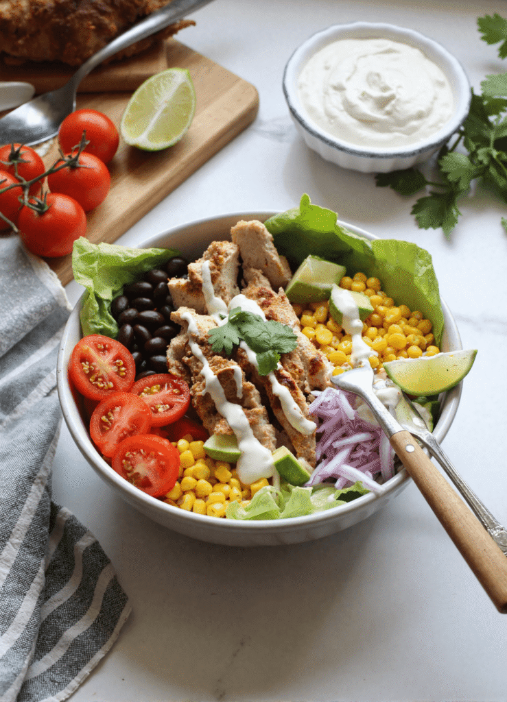 Meal prep Southwest Chicken Salad Bowl with grilled chicken, avocado, red onion, black beans, corn, and lime on a white table.