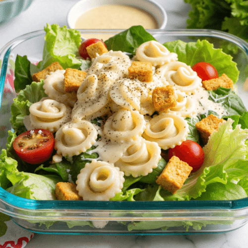 Glass container of tortellini Caesar salad with romaine, cherry tomatoes, croutons, creamy Caesar dressing, and fresh black pepper.
