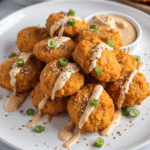 Crispy salmon bites drizzled with bang bang sauce and sprinkled with green onions and sesame seeds on a white plate.