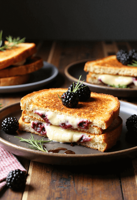 Sliced blackberry balsamic and brie grilled cheese sandwich on a dark ceramic plate, topped with fresh blackberries and garnished with rosemary.