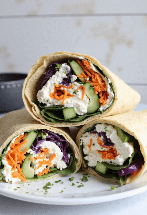 Three high-protein cottage cheese wraps sliced in half and stacked on a plate, filled with spinach, red cabbage, cucumbers, shredded carrots, and creamy cottage cheese.