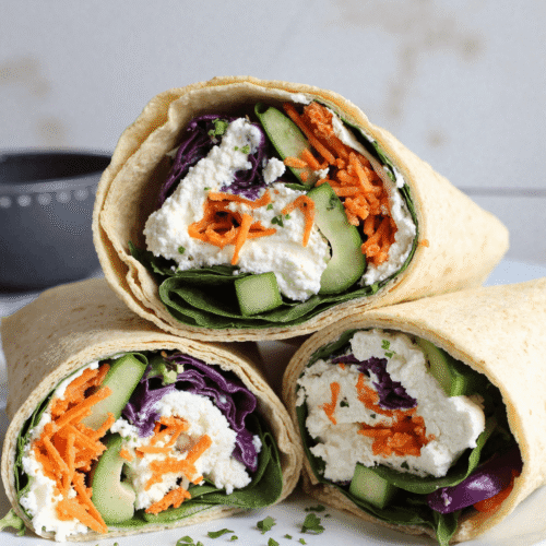 Three high-protein cottage cheese wraps sliced in half and stacked on a plate, filled with spinach, red cabbage, cucumbers, shredded carrots, and creamy cottage cheese.