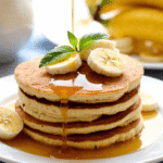 Healthy no-flour banana pancakes stacked with banana slices and syrup drizzle on white plate.