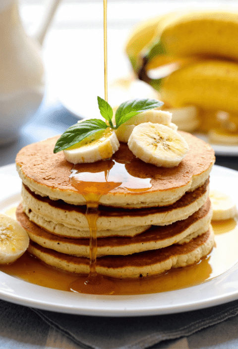 Healthy no-flour banana pancakes stacked with banana slices and syrup drizzle on white plate.