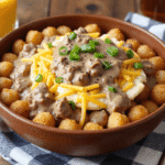 Crispy tater tots topped with fluffy scrambled eggs, rich sausage gravy, shredded cheddar cheese, and sliced green onions in a brown bowl.