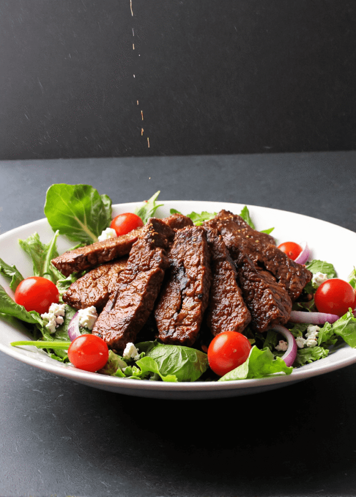 Sliced steak salad on a white plate with cherry tomatoes, red onions, greens, and balsamic vinaigrette being drizzled.